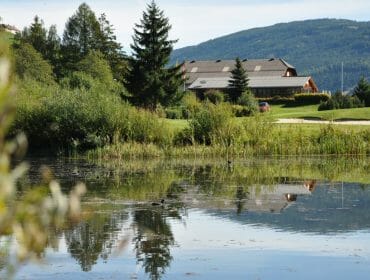 A large house with a sloping roof is reflected in a calm pond surrounded by trees and greenery.