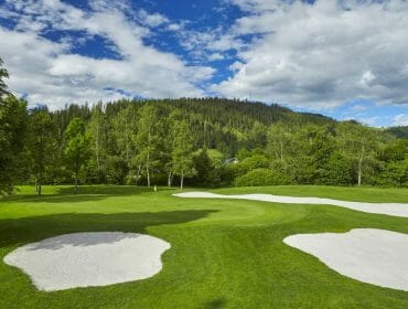 A sunny golf course with sand bunkers, green grass, and trees, set against a forested hillside and blue sky.