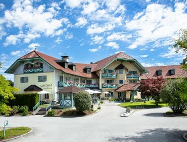 A large alpine-style hotel with balconies, surrounded by greenery and trees under a blue sky with clouds.
