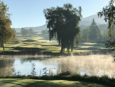 Morning mist rises over a pond with trees and a scenic golf course in the background.