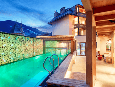A modern outdoor swimming pool with glowing lights next to a cosy mountain lodge at dusk.