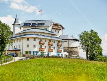 White castle-like hotel with balconies on a grassy hill under a blue sky, with trees and Swiss flag visible.