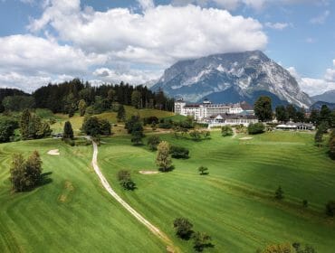 A golf course with a hotel and mountains in the background under a partly cloudy sky.