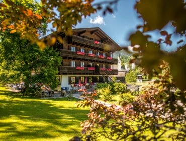 A traditional chalet with red flower boxes, surrounded by green trees and lawn on a sunny day.