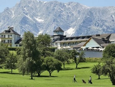 Elegant hotel with golf course in the foreground, set against large, rocky mountains and clear blue sky.