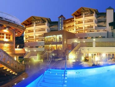 A brightly lit hotel and pool at dusk with alpine architecture and a snowy rooftop.