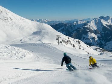 Two people ski down a groomed, snowy mountain slope with chairlifts and mountains in the background.