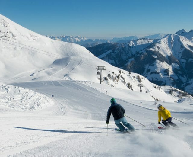 Two people ski down a groomed, snowy mountain slope with chairlifts and mountains in the background.