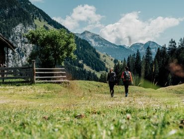 Two people with rucksacks walk on a grassy path towards mountains under a blue sky with clouds.