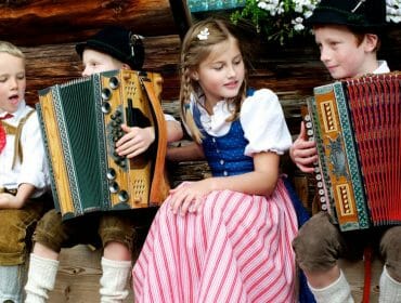 Children in traditional Austrian clothing play accordions and sit together, smiling and talking outdoors.