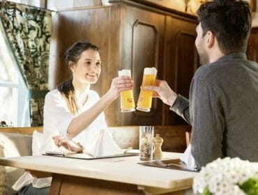 Two people sit at a table in a cosy restaurant, smiling and clinking glasses of beer.