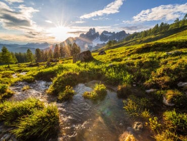 Sunrise over grassy hills and a stream, perfect for walking in the alpine spring among snowy peaks and pines.
