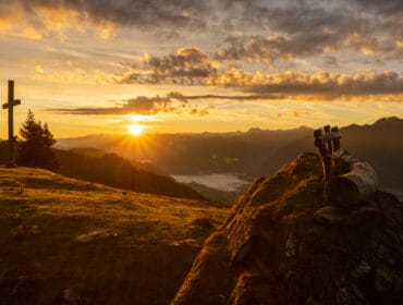 Sonnenaufgang Gamskögerl_© Foto.graf.iererei Moser (2) Sunrise over mountains with a wooden cross, bench, and tree—perfect for Green Spirit: Climate-friendly walking moments.