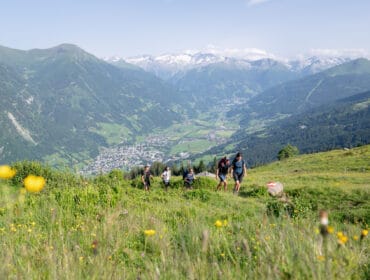 A group of walkers walk up a grassy hill, conquering the whole trail in one piece with mountains and a village behind.