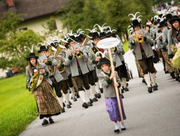 A marching band in traditional Austrian costumes plays instruments in a parade on a rural road.