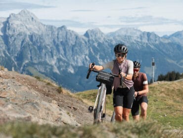 Two cyclists in helmets push their bikes uphill on a gravel mountain trail with tall peaks in the background.