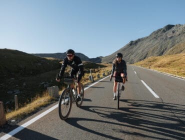 Two cyclists ride on a gravel mountain road under a clear blue sky, surrounded by rocky hills.