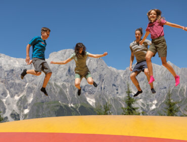 Four children jump on a bouncy castle outdoors with mountains and blue sky in the background.