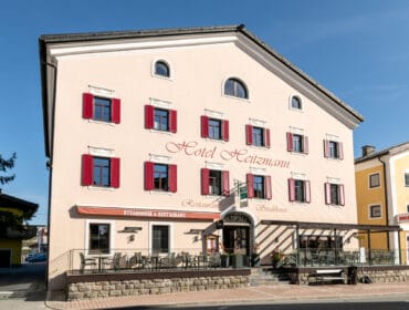 A beige hotel with red shutters, outdoor seating, and Hotel Heitzmann written on the façade.