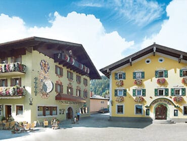 Two traditional alpine buildings decorated with colourful flowers under a bright blue sky with clouds.