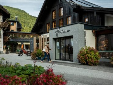 Two people walk past a modern building with flowers in a quaint mountain village setting.