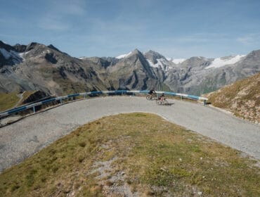 Two cyclists ride on a winding mountain road with snowy peaks in the background under a blue sky.