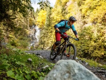 A person mountain biking on a forest trail in Großarltal with a waterfall in the background.