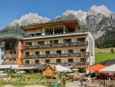 Modern hotel with balconies, a playground, and parasols, set against tall, rocky mountains under a blue sky.