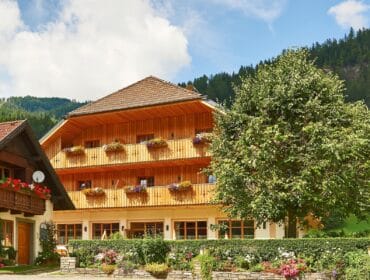 A wooden chalet with flower boxes on balconies, surrounded by greenery and colourful garden blooms.