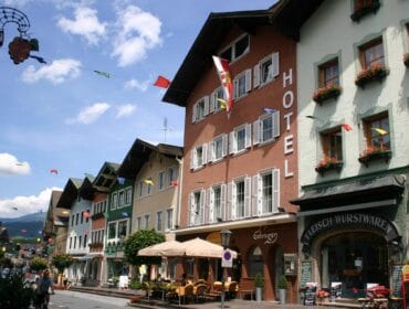 Colourful European street with outdoor café, hotel, and shops, decorated with hanging flags under a blue sky.