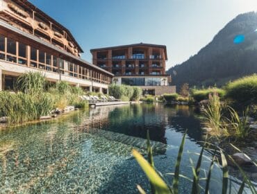 A modern hotel with wooden accents sits beside a clear pond, surrounded by lush greenery and mountains.