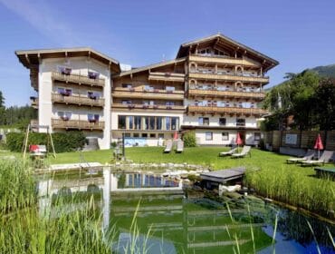 Large alpine-style hotel with balconies, pond, and sun loungers on a grassy lawn under a clear blue sky.