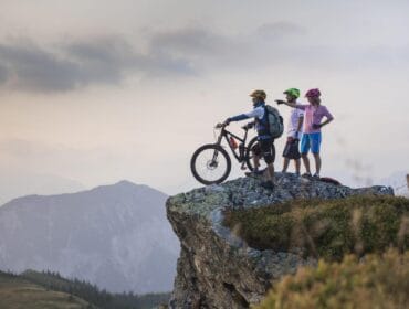 Three cyclists with helmets stand on a rocky cliff, overlooking mountains under a cloudy sky.