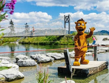 Person in a lion costume stands on a floating platform by a rope bridge over a pond, with people in the background.