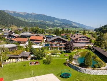 Aerial view of a hotel with gardens, a pond, and mountains in the background under a clear blue sky.