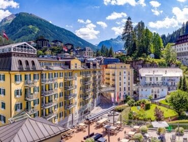 Colourful hotels and buildings surrounded by mountains and greenery under a blue sky in a scenic alpine town.
