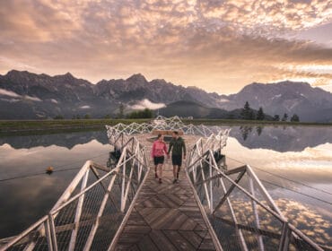 prinzensee_natrun_maria-alm_foto-christian-schartner-42 Two people walk on a modern bridge over calm water with mountains and a dramatic sky in the background.