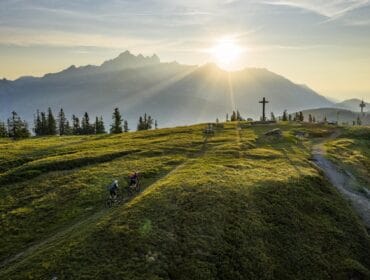 Two cyclists ride up a grassy mountain trail at sunrise in Salzburger Sportwelt, cross and peaks behind them.