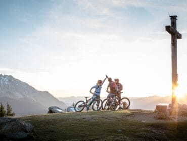 stoneman_taurista_rossbrandcsalzburger_sportwelt Three mountain bikers pause at sunrise near a wooden cross in the Salzburger Sportwelt mountains.