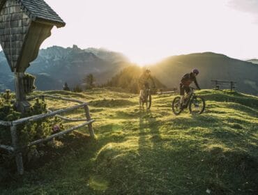 Two cyclists ride on a grassy hill at sunrise in Tennengau, with mountains and wooden fences in the background.