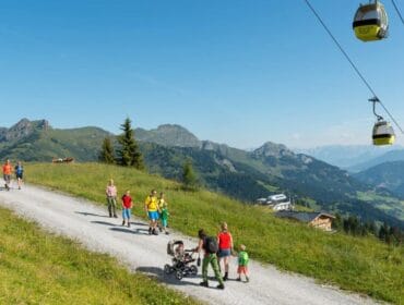 People hiking on a mountain footpath with cable cars overhead and picturesque hills in the background under a blue sky.