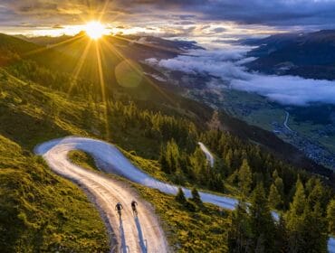 Two cyclists ride at sunrise on a winding Wildkogel Arena mountain road, surrounded by trees and distant valleys.