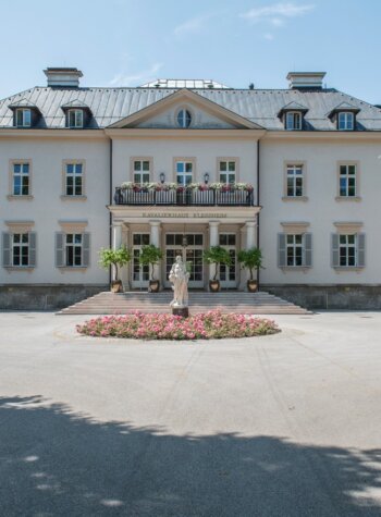 Large white mansion with statue and flowers, seen through ornate gates—explore with SmartGuide audio guide.