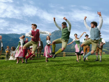People in traditional Austrian clothing dance joyfully on a grassy field with mountains in the background.