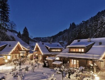 Snow-covered cabins with glowing lights nestled in a forested mountain village at dusk.