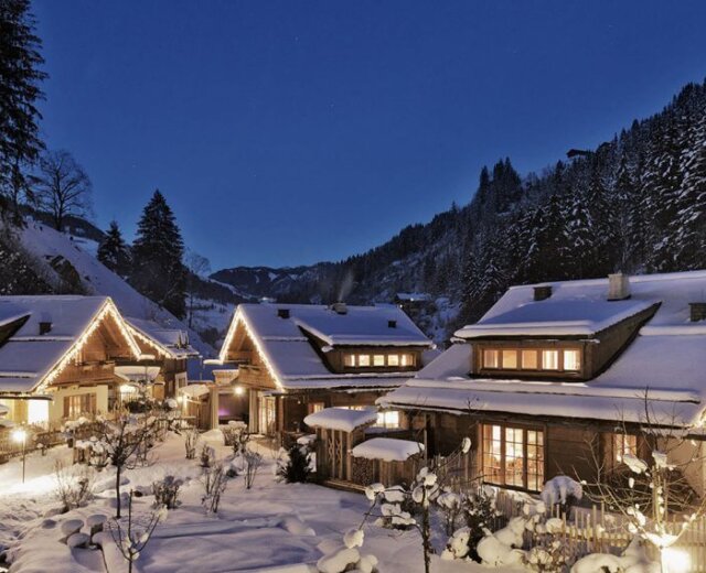 Snow-covered cabins with glowing lights nestled in a forested mountain village at dusk.