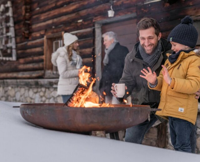 A man and a boy warming their hands by a fire pit outside an Österreich wooden cabin in the snow.