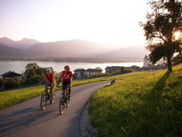 Two cyclists ride the Salzkammergut Cycle Trail by a lake at sunset, with mountains and a village in the background.