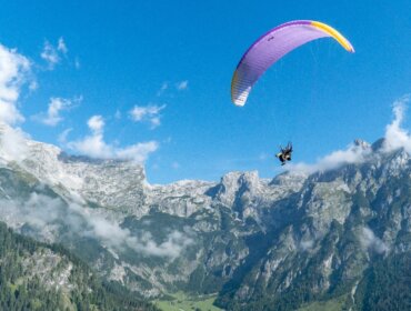 A person paragliding over green mountains with a bright blue sky and a few clouds in the background.