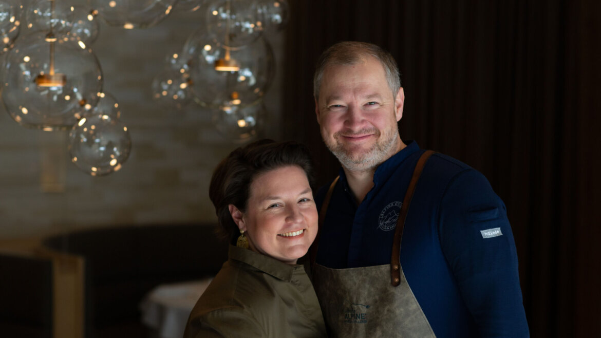 A smiling couple stands together indoors, with modern glass light fittings in the background.